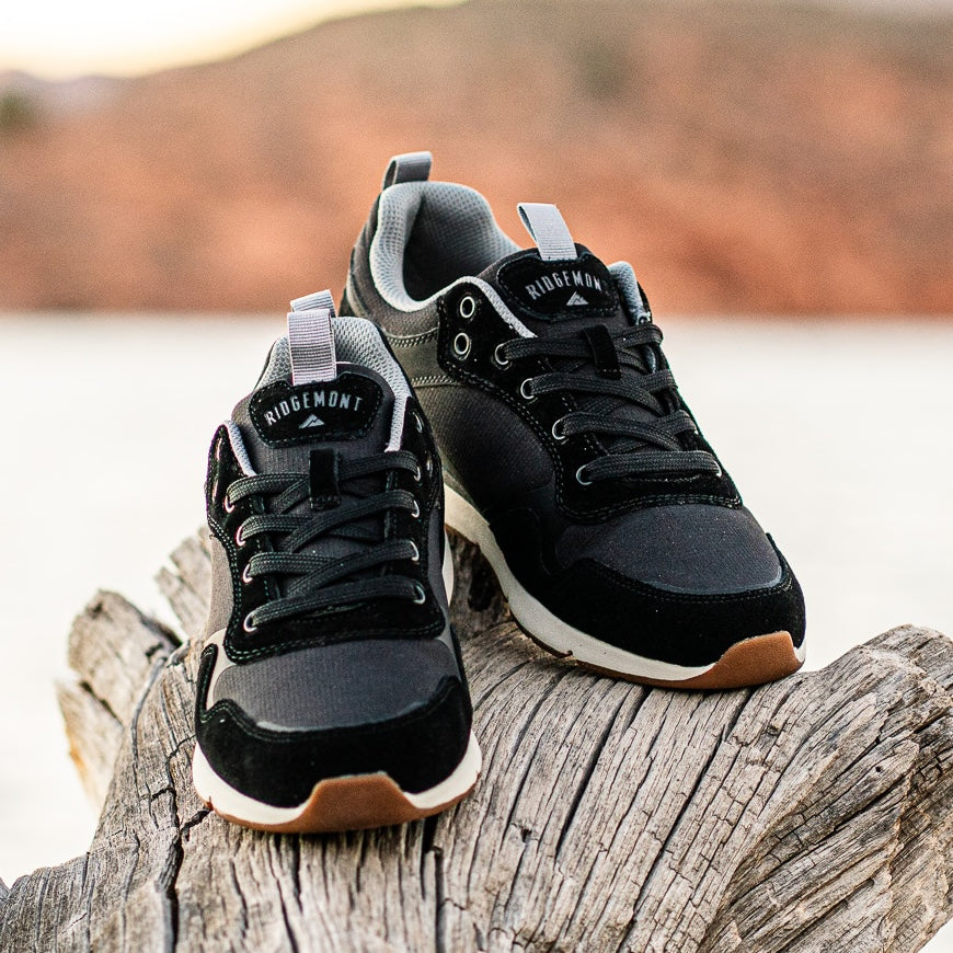 Pair of Ridgemont Escape sneaker in color Black. Shoes displayed outside by river in St. George, Utah, laying on wood. Shoes have black oiled suede, black canvas, black ripstop poly, black laces, white CMEVA midsole and grey inner lining, real rubber gum colored tread with study grip. Outdoor shoe, everyday sneaker, comfortable pair of shoes, Everyday comfortable sneakers, walking shoes.