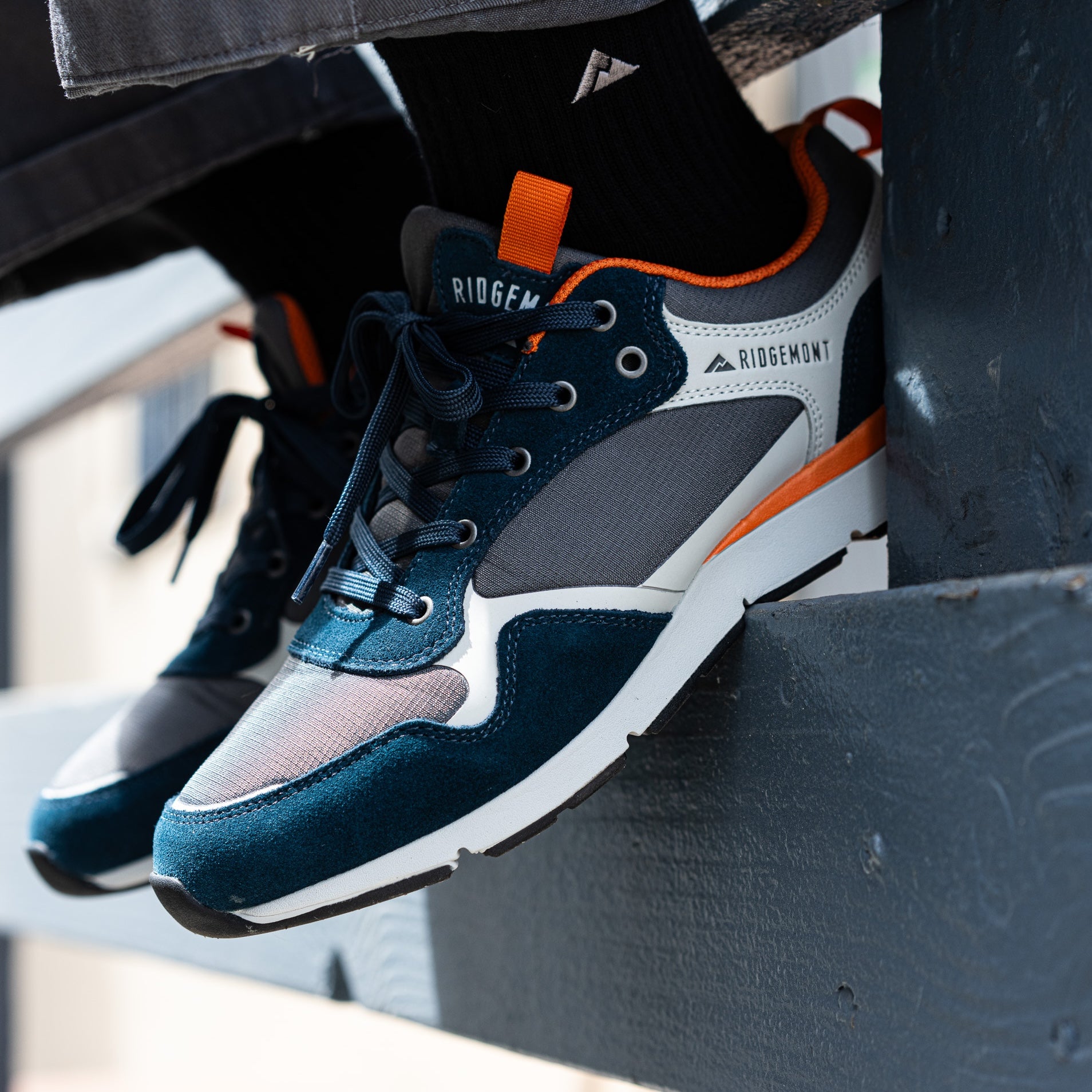A close-up of a person wearing a pair of Ridgemont Escape sneakers in Navy Charcoal, with one shoe resting on a dark metal surface. The sneakers feature a dark teal suede and mesh upper with gray panels, white midsoles, and bright orange accents.