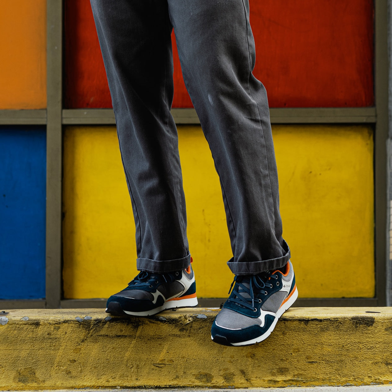 A person wearing dark gray pants and pair Escape Navy Charcoal sneakers stands on a weathered yellow curb in front of a colorful block-patterned wall.