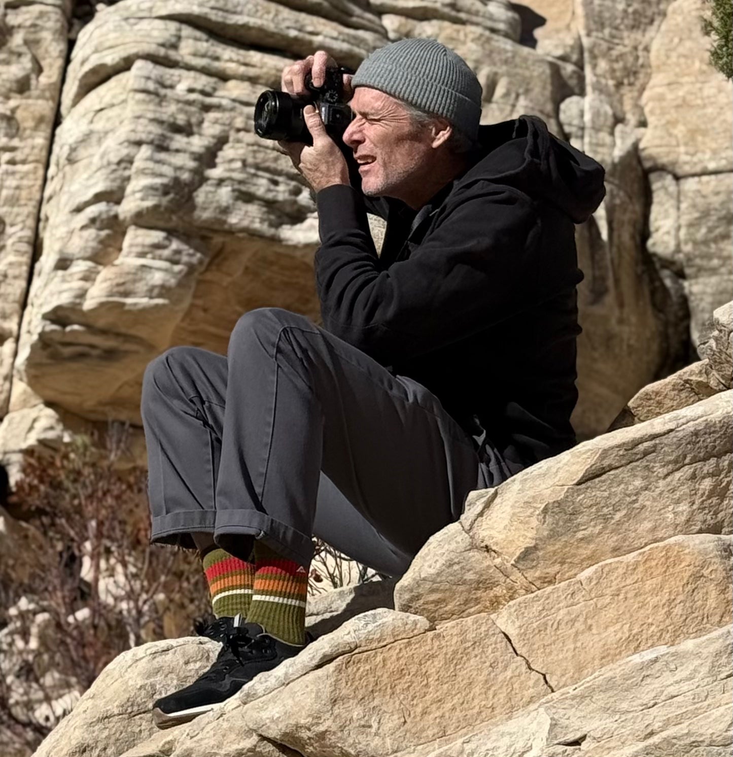 Photographer is wearing Ridgemont Escape Sneaker in color Black and Ridgemont Trailblazer Crew sock in color Olive. Photographer is sitting on rock face. Black Escape sneaker shoe has white CMEVA midsole, real rubber tread in color gum, black ripstop poly, black oiled suede, black canvas, black laces, grey inner lining. Everyday shoes, walking shoes, comfort, comfortable, low top sneaker.