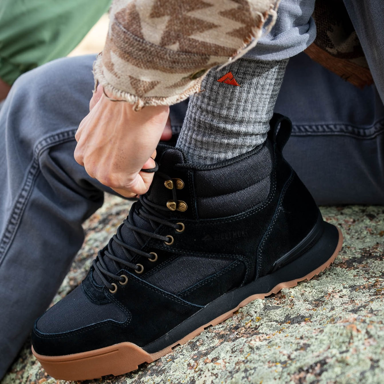 A close-up of a person wearing a black Monty boot with black laces, gold-tone eyelets, and a gum sole, sitting on a rock outdoors while wearing a Ridgemont Grey Staple Sock