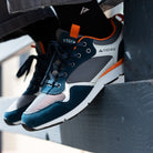 A close-up of a person wearing a pair of Ridgemont Escape sneakers in Navy Charcoal, with one shoe resting on a dark metal surface. The sneakers feature a dark teal suede and mesh upper with gray panels, white midsoles, and bright orange accents.