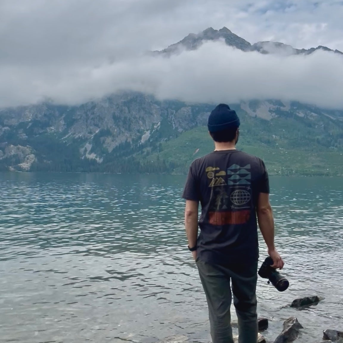 Jerrick Romero in the Ridgemont Remod T-shirt with camera in hand standing in front of a lake