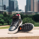 Pair of Ridgemont Outback boot in colors Black Grey with black rubber tread and lugged sole (outsole) and white foxing tape. Side view of shoe in city landscape. Outdoors. In Austin, Texas.