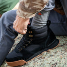 A close-up of a person wearing a black Monty boot with black laces, gold-tone eyelets, and a gum sole, sitting on a rock outdoors while wearing a Ridgemont Grey Staple Sock