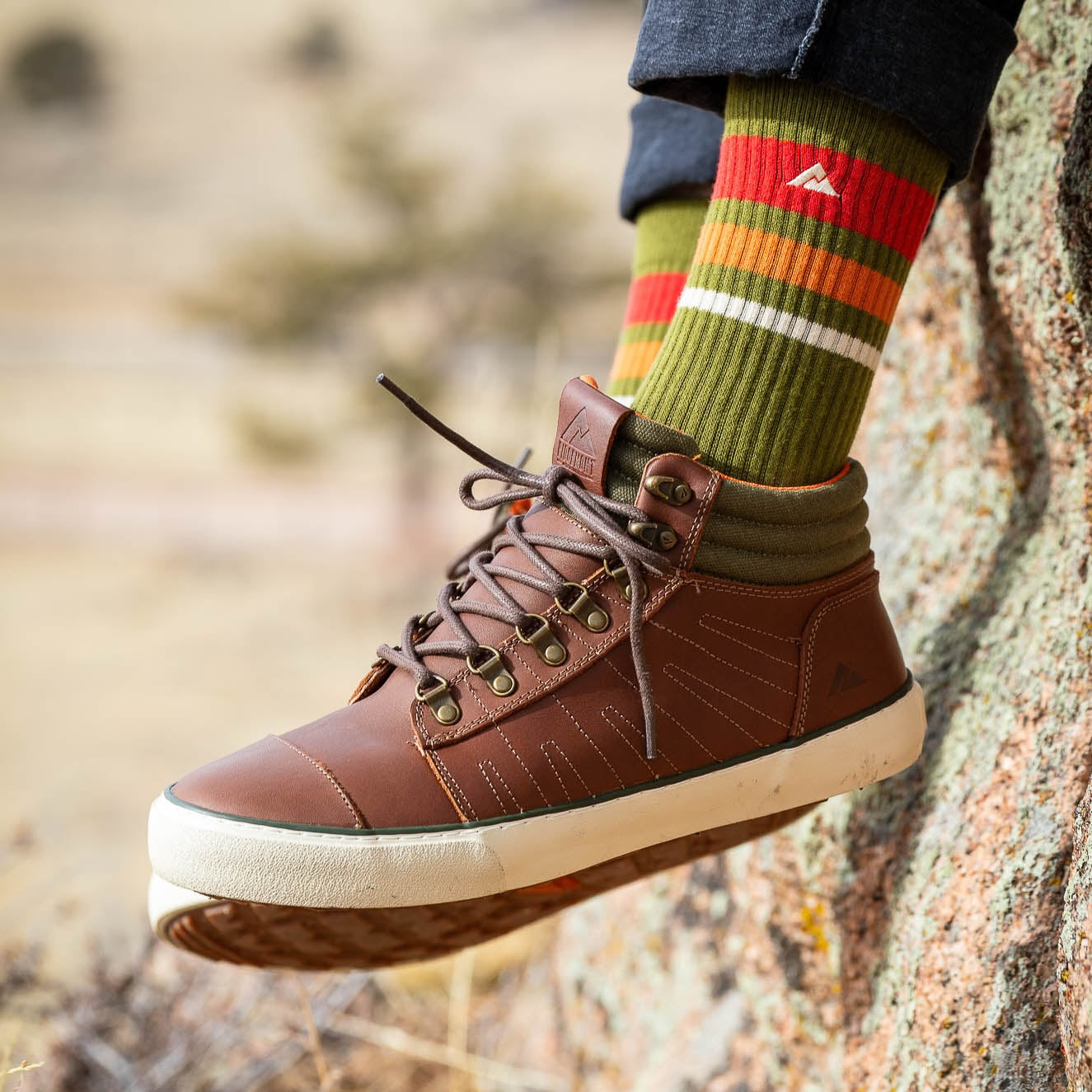A person's foot wearing Outback boots in Brown Olive leather high-top shoe with a thick cream sole, hanging against a rock outdoors. The person is also wearing dark jeans and Ridgemont Olive Trailblazer crew socks with red, orange, and white stripes and a white Ridgemont  logo.